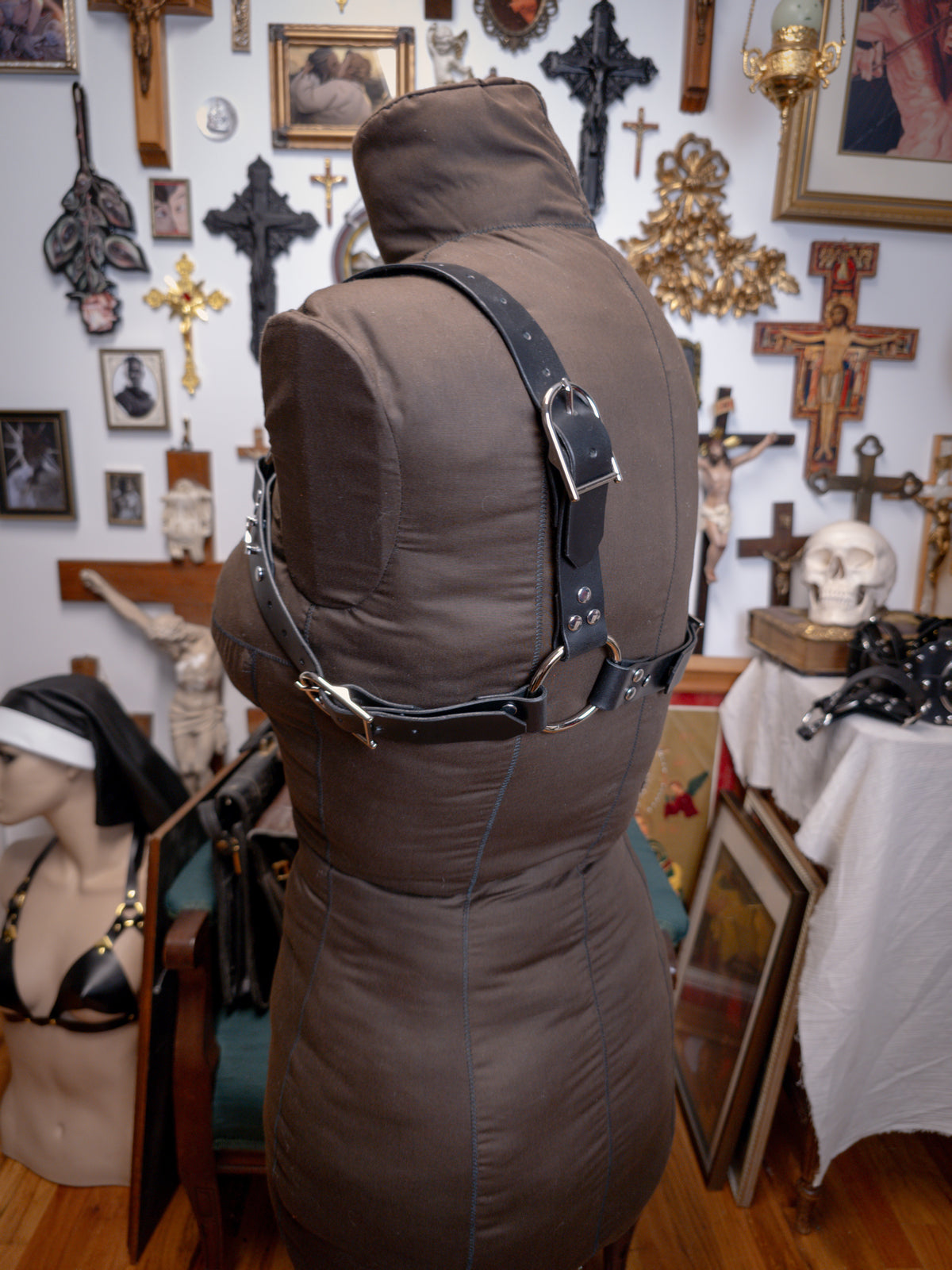 side back view of black leather harness with crosses on a brown fabric mannequin against a gothic background