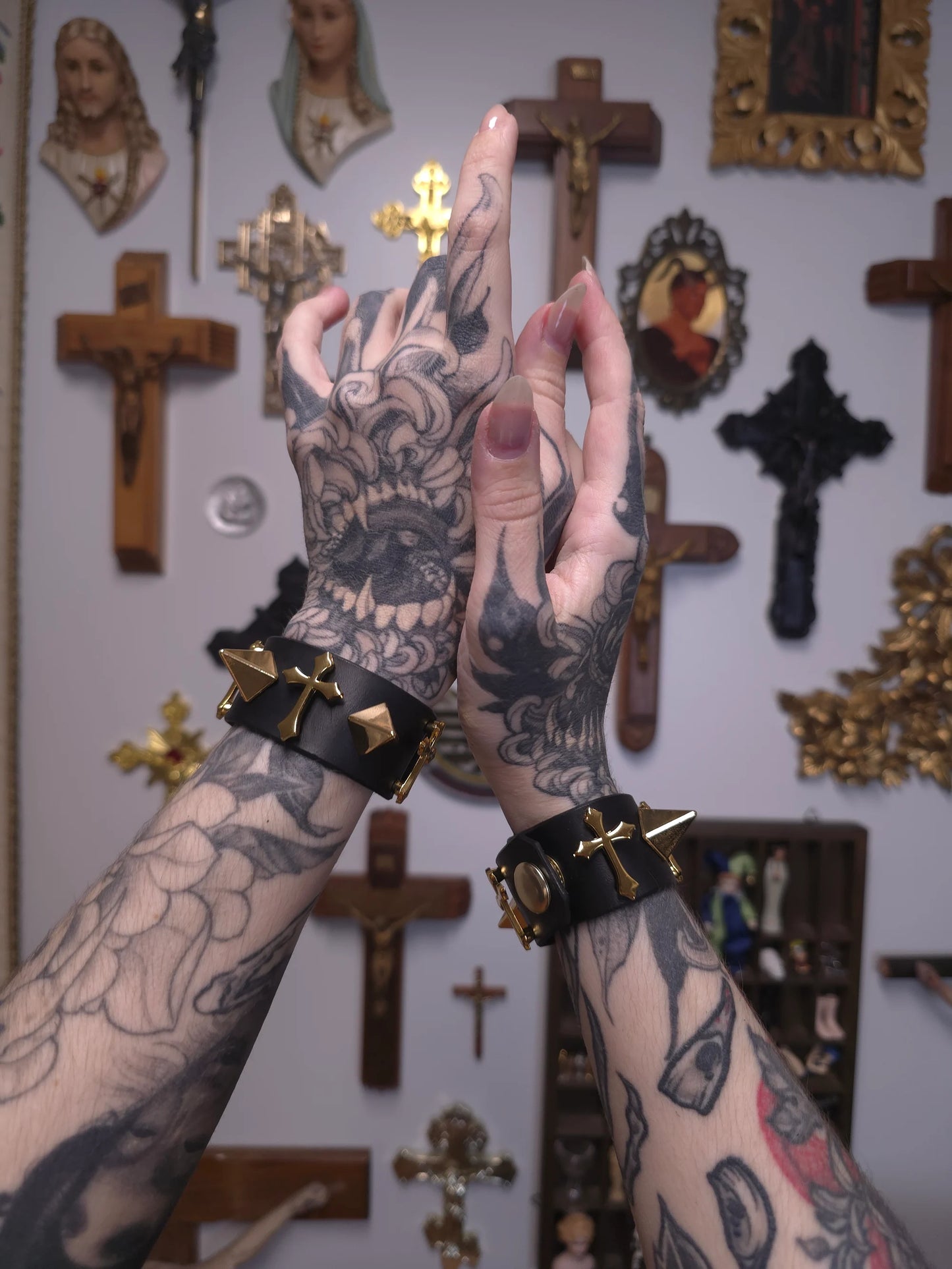 Tattooed hands with black leather bracelets in front of a wall with crosses and religious statues.