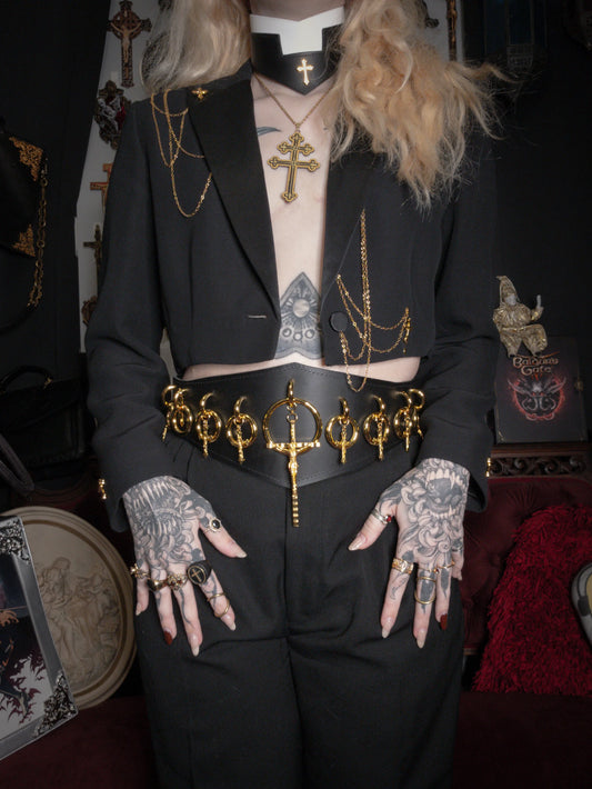 Person wearing a black outfit with gold accessories and tattoos, standing in a dark room.