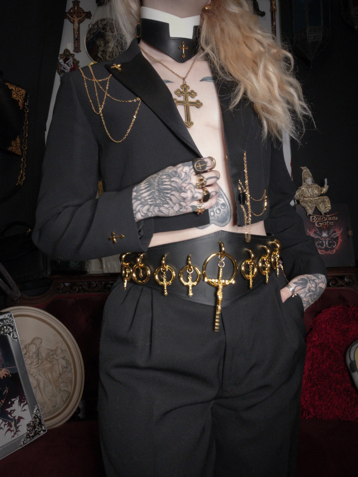 Person wearing a black outfit with gold accents and tattoos, standing in a dark room.