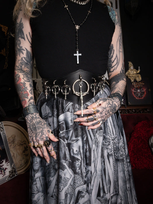 Person wearing a black top with a cross pendant and a detailed skirt, surrounded by tattoos and jewelry.
