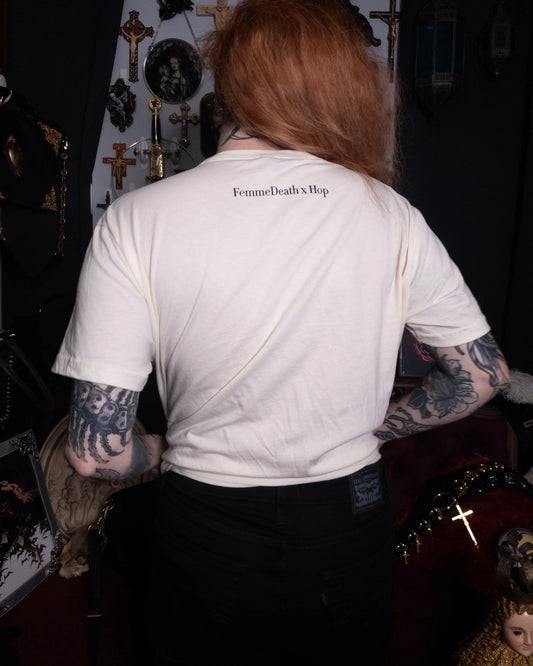 Person wearing a cream t-shirt with 'FemmeDeath x Hop' text, standing in a dark room with crosses and religious symbols.