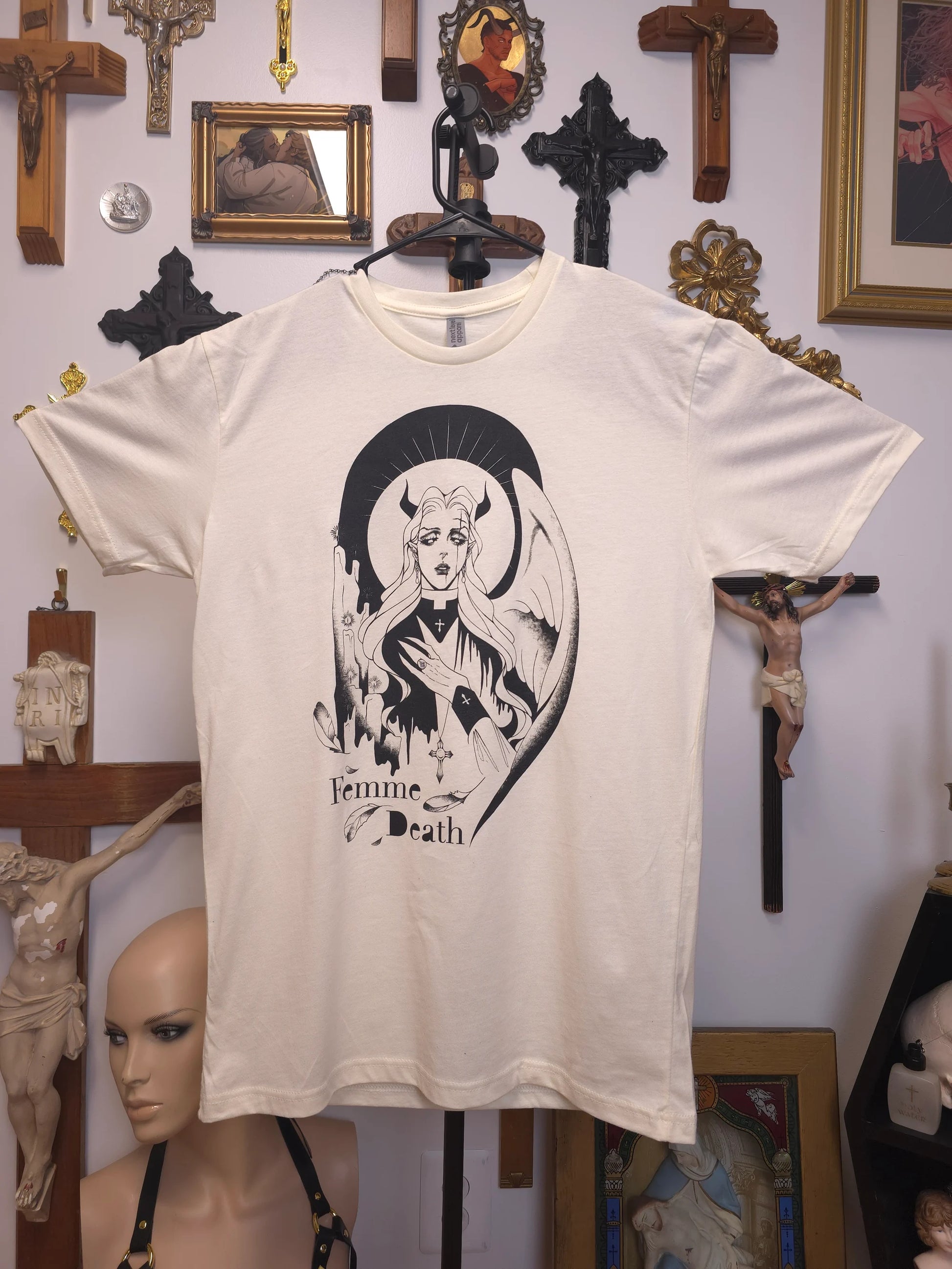 White t-shirt with 'Femme Death' design on a hanger against a wall with religious-themed decorations.