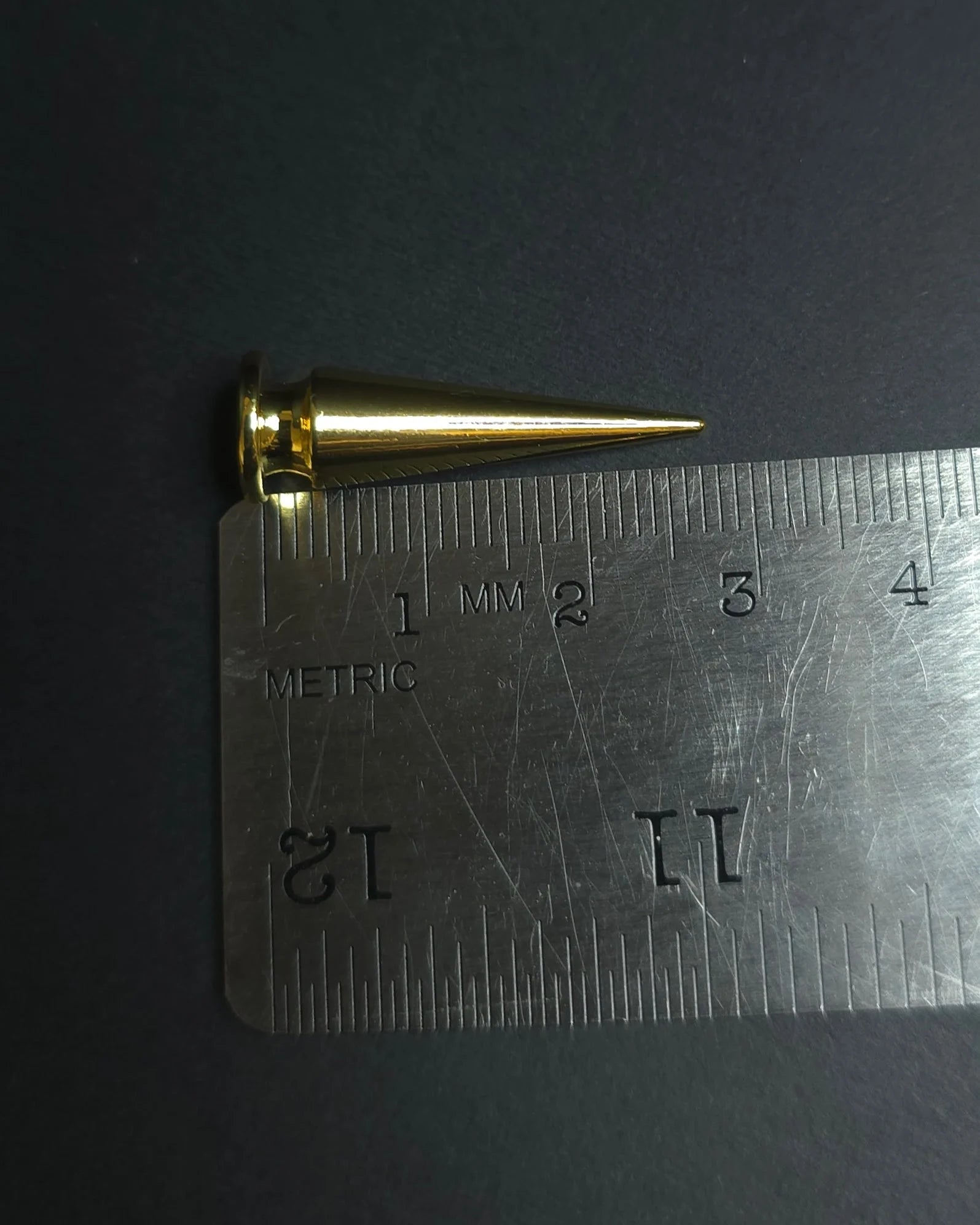 Gold cone-shaped spike next to a metal ruler on a dark background