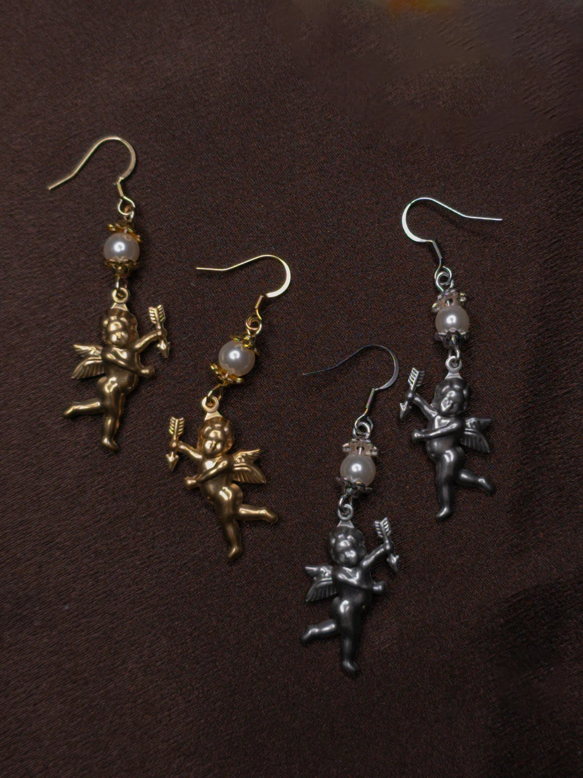 Two pairs of angel-shaped earrings in gold and silver on a dark brown background.