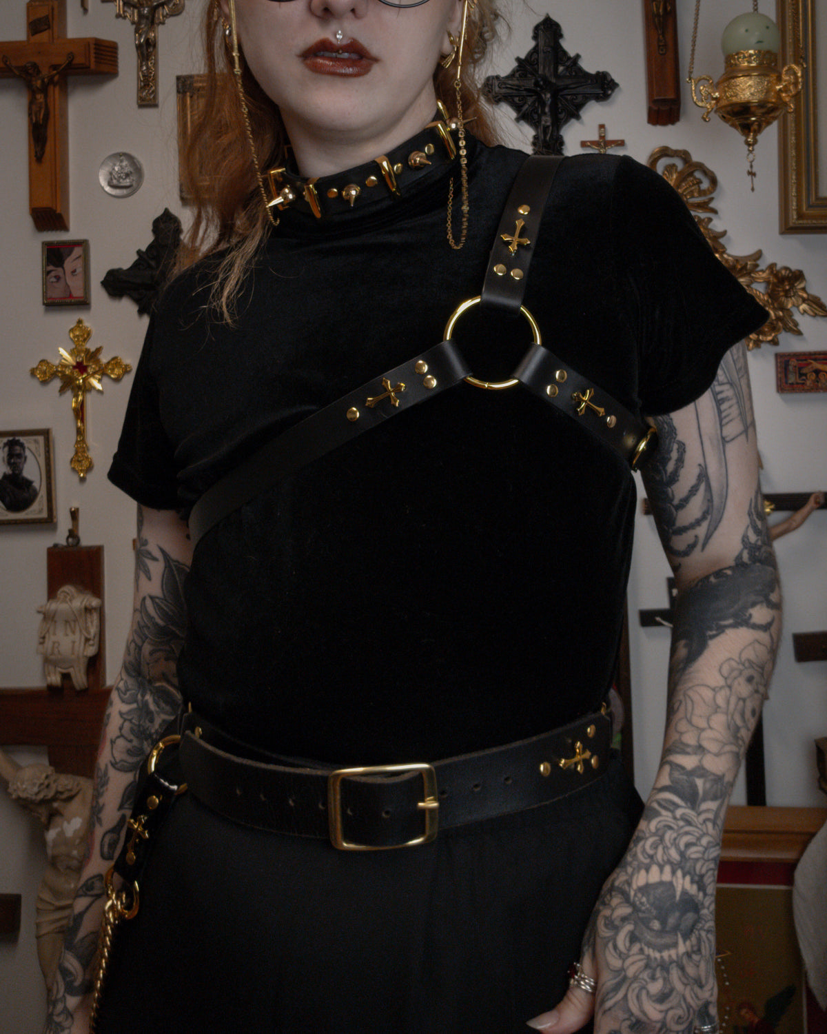 a person wearing a black velvet top with a black leather asymmetrical harness on top, golden spiked collar and golden buckle waist belt, against a gothic background