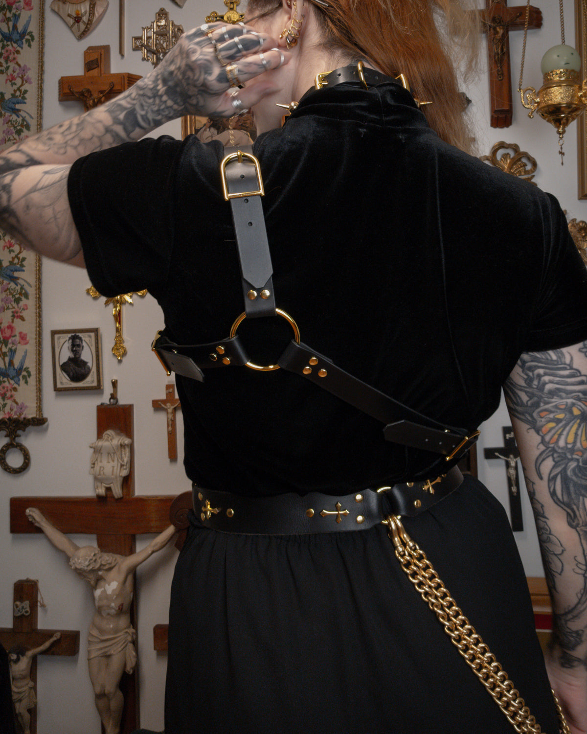 back view of a person wearing a black velvet top with a black leather asymmetrical harness on top, golden spiked collar and golden buckle waist belt, against a gothic background