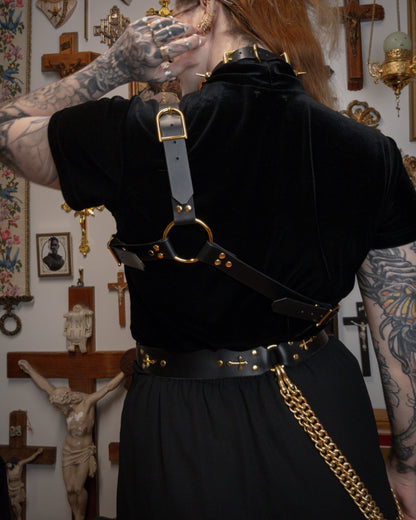 back view of a person wearing a black velvet top with a black leather asymmetrical harness on top, golden spiked collar and golden buckle waist belt, against a gothic background