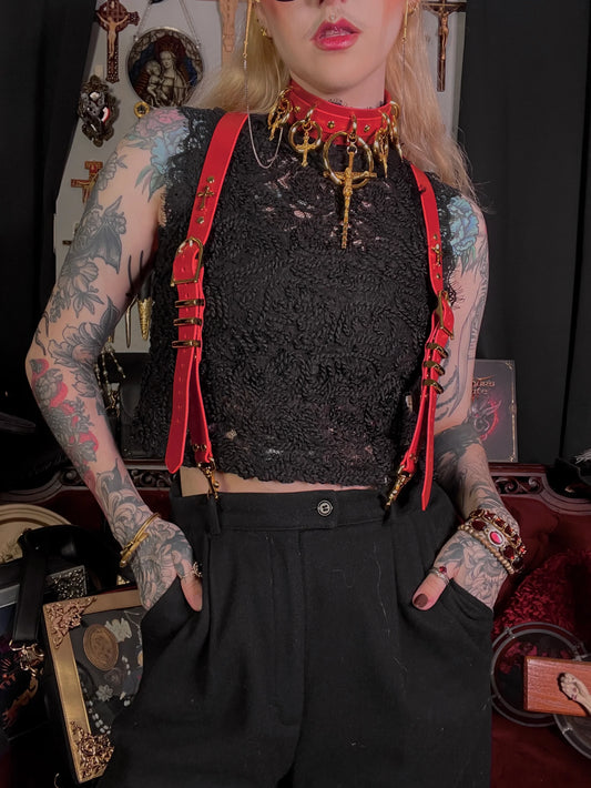 Person wearing a black lace top with red suspenders and tattoos, standing against a dark background.