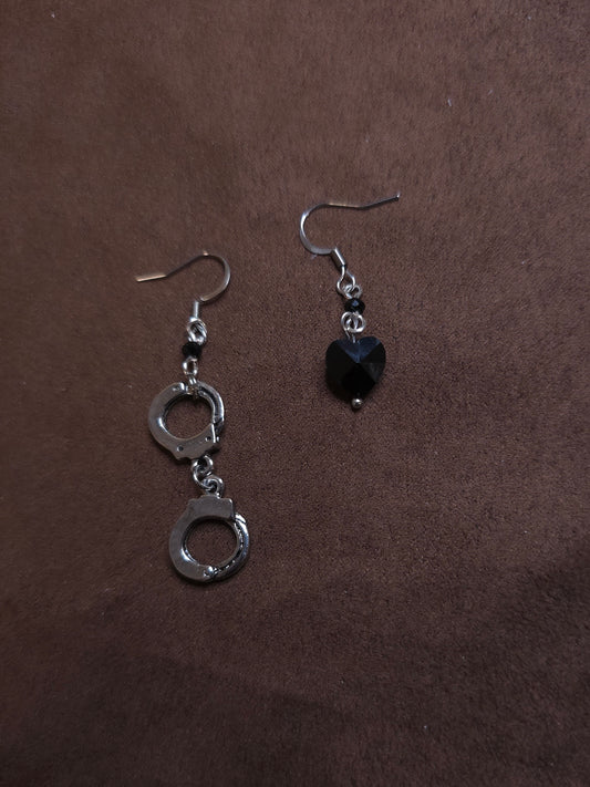 Pair of silver handcuff-shaped earrings with a black heart charm on a brown background